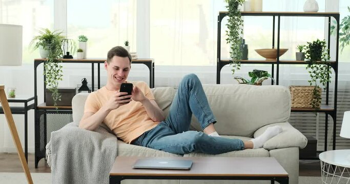 Happy man seated on a cozy couch in the living room, fully immersed in his smartphone. A warm smile graces his face as he engages with the content on his device, finding joy in what he sees.