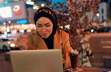 A hijab woman enjoying a sweet dessert at a coffe while working on her laptop during the nighttime in a bustling urban setting.