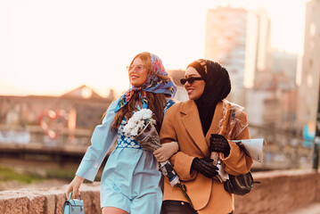 Fototapeta na wymiar Couple woman one wearing a hijab and a modern yet traditional dress, and the other in a blue dress and scarf, walking together through the city at sunset. One carries a bouquet and bread, while the