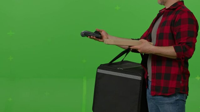 Caucasian male courier passing a box of pizza on green screen chroma key background. Man hold wireless bank payment POS terminal to process acquire credit card payment. Copy space mock up advertising
