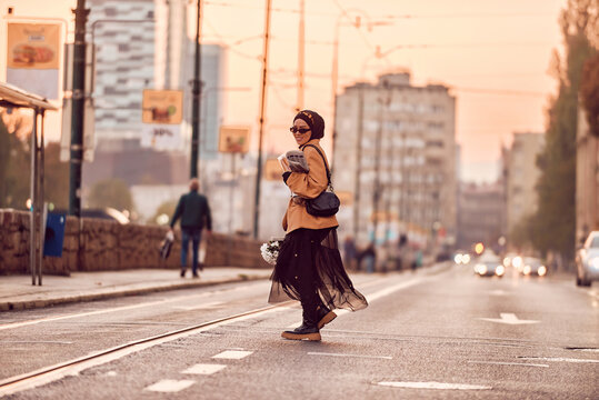 A hijab woman, donning a luxurious vintage French outfit, as she walks through the city streets at sunset, carrying bread, a bouquet of flowers, and newspapers in her hand, evoking a captivating blend