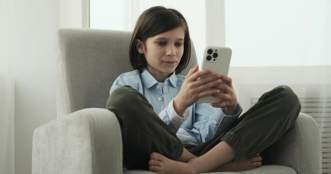 A focused Caucasian boy seated in an armchair, deeply engrossed in his phone. With his undivided attention, he navigates through apps, messages, or perhaps explores a captivating digital world.