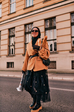 A hijab woman, donning a luxurious vintage French outfit, as she walks through the city streets at sunset, carrying bread, a bouquet of flowers, and newspapers in her hand, evoking a captivating blend