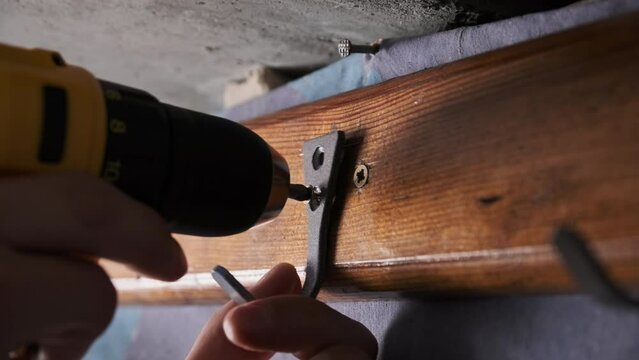 Craftsman screwing a self-tapping screw into a board using an electric screwdriver. Screwing clothes hook to wooden plank on a wall using a power drill, close-up. Concept handmade, do it yourself