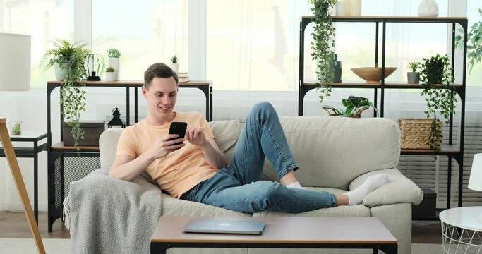 Joyful Caucasian man as he engages in lively conversations and bursts into laughter while using his phone on the sofa. With a bright smile on his face and an unmistakable twinkle in his eyes.