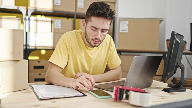 Young hispanic man ecommerce business worker using smartphone and laptop writing on document at office