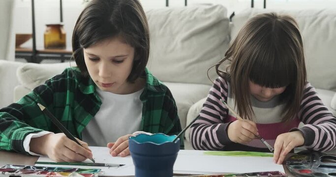 Siblings seated near a table on the floor at living room, they dive into their creative pursuits, skillfully using pencils, paintbrushes, and watercolors.