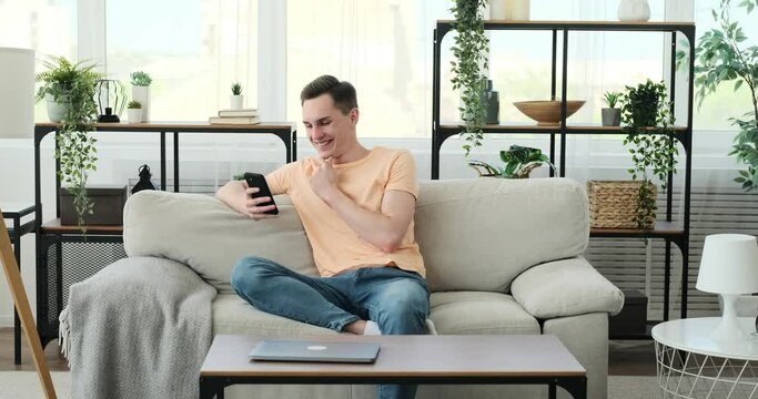 Man seated on the couch, his face adorned with a genuine smile, and his attention captivated by the phone in his hands. Completely at ease, he indulges in the digital realm, letting go of any worries.