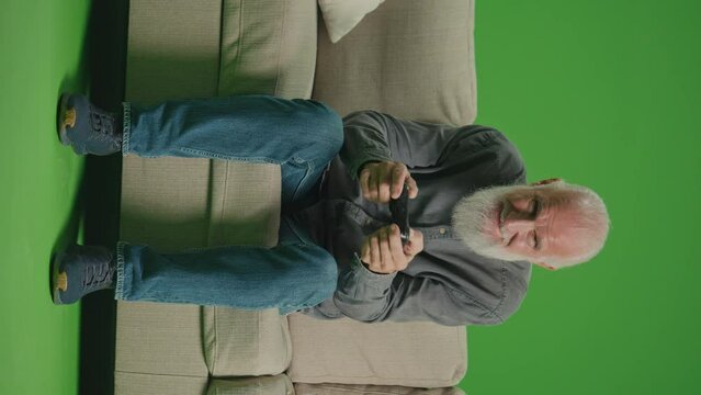 Vertical View.Green Screen. An Old Man With a Gray Beard Plays Computer Games With a Joystick. An Elderly Man Plays PlayStation and Wins.Tech Nostalgia and Generational Divide.