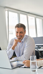 Fototapeta premium Happy professional mid aged mature Latin executive manager working in office with female colleague, talking and looking at laptop computer sitting at table workplace. Vertical authentic shot.