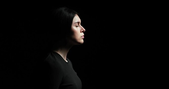 Woman standing in profile on a black background, breathing deeply and dramatically lifting her head to gaze upwards. With a serious expression and a thoughtful demeanor.