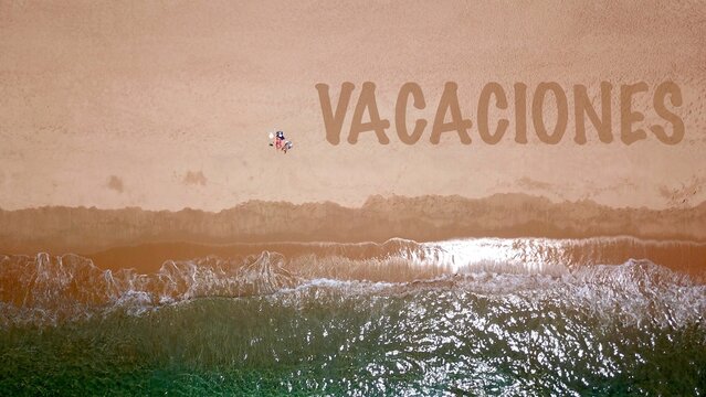 Vista a&eacute;rea de una hermosa playa de arena junto al mar azul, donde dos vacacionistas yacen y toman el sol, junto a ellos las letras "VACACIONES" en la arena