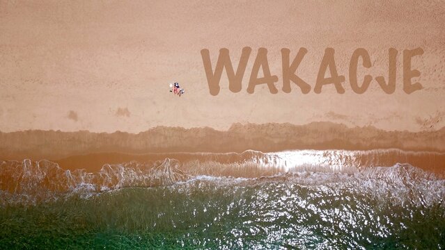 Widok z lotu ptaka na piękną piaszczystą plażę nad błękitnym morzem, gdzie leży i opala się dw&oacute;ch wczasowicz&oacute;w, obok nich napis "wakacje" na piasku