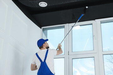 Worker in uniform painting ceiling with roller indoors