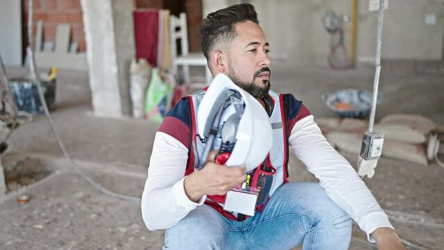 Young latin man builder sitting on floor using hardhat as a hand fan at construction site