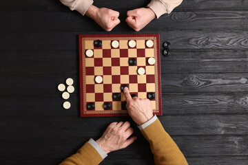 Senior man playing checkers with angry partner at black wooden table, top view