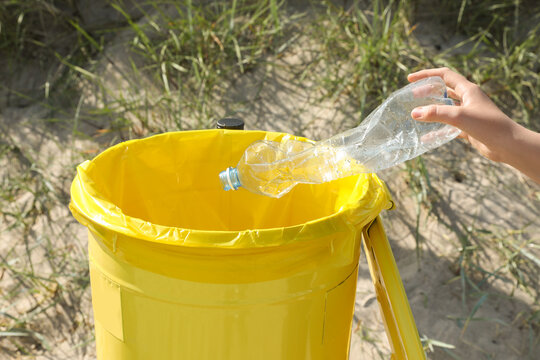 Woman Throwing Plastic Bottle In Yellow Bin On Beach, Closeup. Recycling Concept