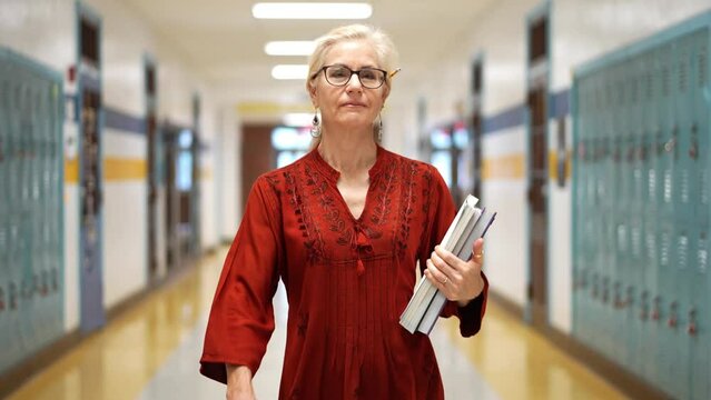 Closeup slow motion portrait of pretty mature woman teacher holding books walking towards camera down an empty school hallway.