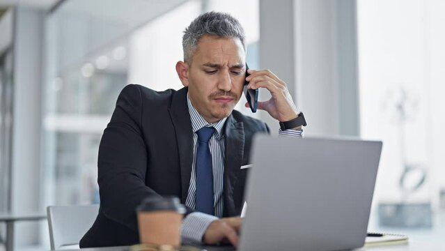 Young hispanic man business worker talking on smartphone using laptop at office
