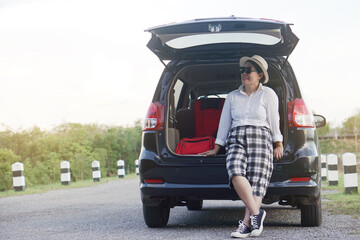 Happy middle aged Asian woman traveler sits at the trunk of car. Concept, relax time. Vacation or holiday trip by private vehicle. Transportation. Give time for yourself.   