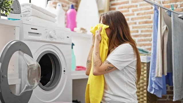 Young beautiful hispanic woman smelling clean clothes doing thumb up gesture at laundry room