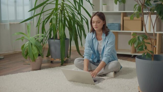 Smiling Young Woman Sitting at Laptop on the Floor Among Flowers. Plants Improve Mood and Well-being, Woman Works at Home, Woman Studying at Home with Laptop, Woman is Preparing a Project on Laptop
