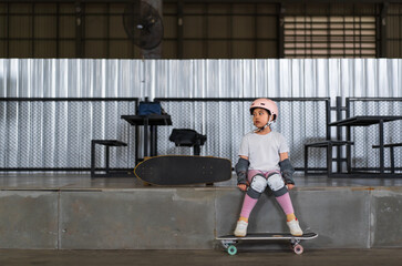 asian child skater or kid girl sitting relax at skatepark with skateboard or surf skate to people extreme sports in indoor surfskate skate park and wears helmet elbow wrist knee guard for body safety