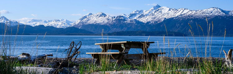 Fototapeta na wymiar Wood picnic table on the on beach of Katchemak Bay on a sunny summer day, mountain range in the background, Homer, AK 