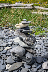 Beach rocks balanced in a stack on a rocky beach with grass and driftwood in the background
