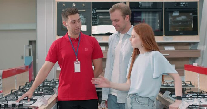 Salesman demonstrating new stove to attractive couple in kitchen furniture shop. young husband and wife choosing electric oven consulting manager in modern showroom Slow motion
