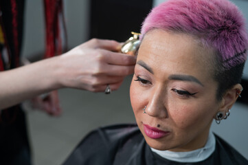 The hairdresser shaves the temple of a female client. Asian woman with short pink hair in barbershop.