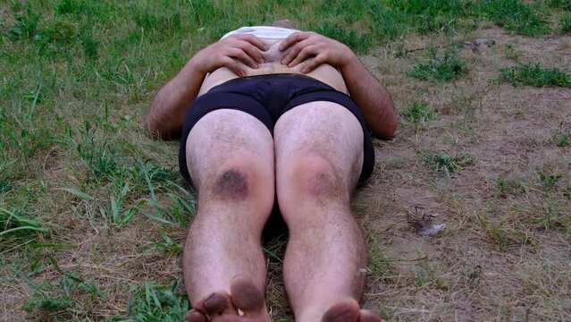 Barefoot obese man wearing white shirt and only briefs lying on the ground while massaging his hairy stomach.