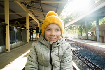 Portrait of a smiling boy in yellow hat and grey down jacket at the train station under sunlight.