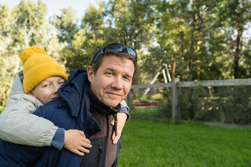 Fototapeta premium Father giving his son a piggyback ride outdoors. Lifestyle portrait, copy space. Autumn.