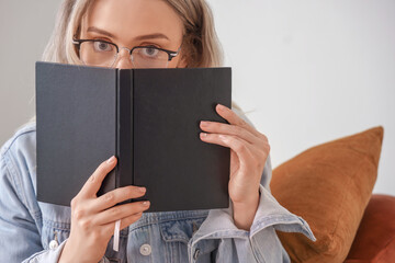 Obraz premium Young woman in glasses peeking over black book, reading and learning concept, education and knowledge acquisition