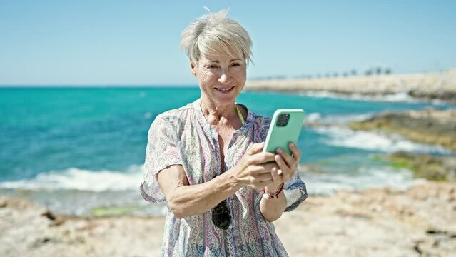 Middle age blonde woman tourist smiling confident using smartphone at the beach