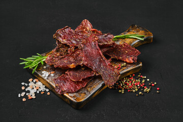Dried beef jerky slices on wooden board with salt and pepper.
