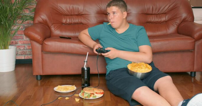 plump overweight teenager boy playing video game, eating french fries, sitting resting on floor, Slow motion, leisure lifestyle free spare time hobby interests