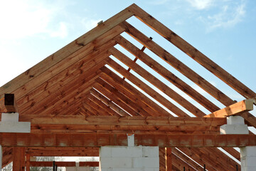 A timber roof truss in a house under construction, walls made of autoclaved aerated concrete blocks, rough window openings, a reinforced brick lintel, a scaffolding, blue sky in the background