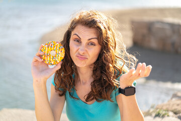 Young sport woman holding a donut at outdoors making doubts gesture while lifting the shoulders