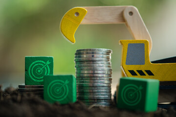 Stacked coins in soil with target icons on a green wood block and loader car. Money growth concept....