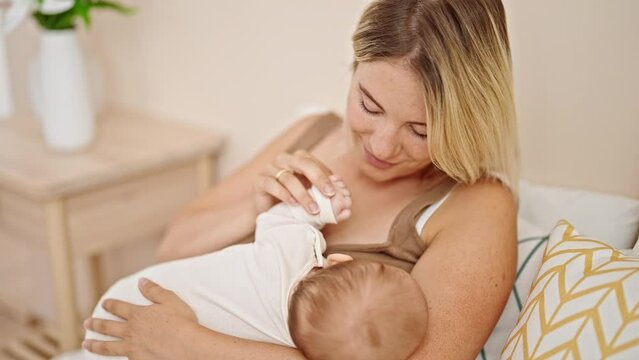 Mother and daughter sitting on bed breastfeeding baby kissing hand at bedroom