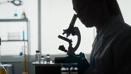 Side view of a dark silhouette of a female scientist looking under a microscope, doing an analysis...