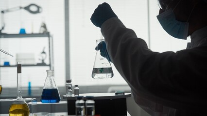 Dark silhouette of a male scientist conducting a scientific experiment close up. A man using a pipette will add a green reagent to a glass flask with a clear liquid.
