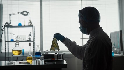 A male scientist holds an Erlenmeyer flask with a plant inside and examines it. Dark silhouette of a scientist with a flask in his hands in the laboratory. Scientific experiment.