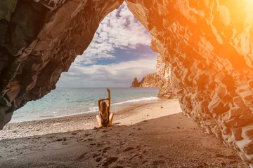 Gordijnen Woman travel sea. View of a woman in a black swimsuit from a sea cave Attractive woman enjoying the sea air sits on the beach and looks at the sea. Behind her are rocks and the sea © svetograph