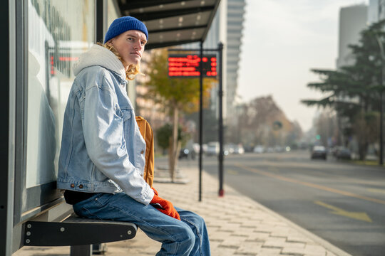 Blond guy student waiting transport very long time in morning. Man on bus stop in sunny weather. Blonde hair millennial man on bus stop in city. Modern urban transport for everyone