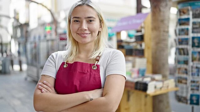 Young beautiful hispanic woman wearing apron standing with arms crossed gesture at newspaper store