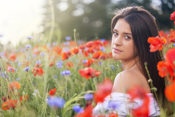Naklejka premium Closeup side profile view of cool brunette girl exposing her shoulder sitting in red poppies and blue cornflowers meadow. 