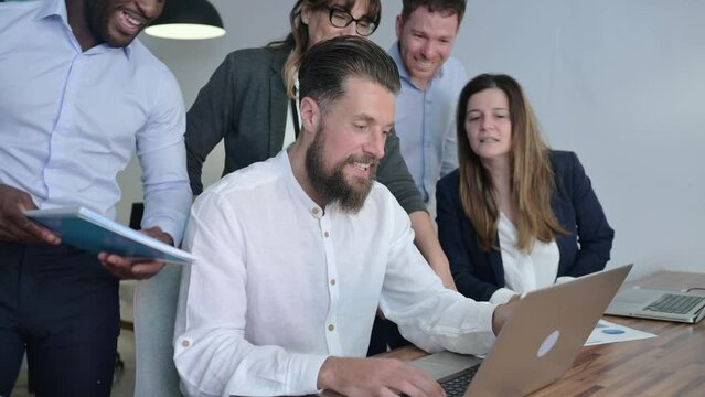 Group of diverse colleagues in formal clothes discussing project details while standing and seating near table with laptop in office